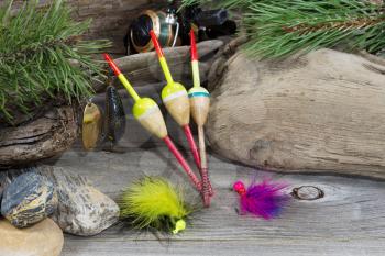 Horizontal image of fishing equipment consisting of floats, reel, lures, and feathered flies resting against aged driftwood, evergreen tree branches and rocks 