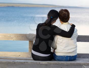 Closeup of mother with mature daughter holding her while sitting on wooden bench looking outward at the lake

