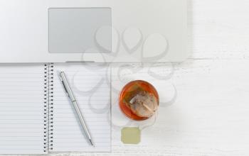 Top view angle shot of desktop with notepad, pen, laptop computer and glass of tea with tea bag inside.  Horizontal format. 