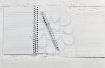 Top view angle shot of white wooden desktop with notepad and silver pen. Horizontal format. 
