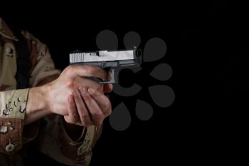 Close up horizontal image of pistol, pointing sideways into darkness, with armed male soldier in background. Focus on side part of weapon. 