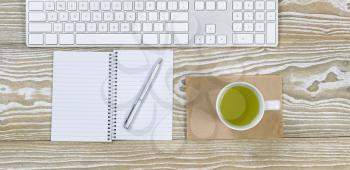 Top view shot of an old white desktop with keyboard, green tea in cup, notepad and pen in horizontal format.