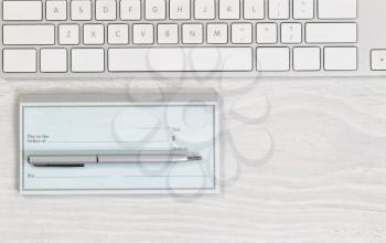 Image of partial keyboard with blank checkbook and silver pen on white desktop. Layout in horizontal format.