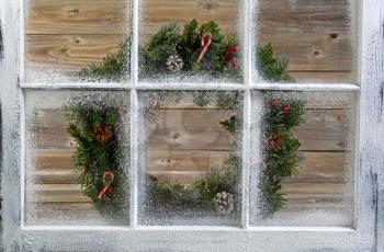 Snow covered window with decorative Christmas wreath on window with rustic wood in background. 