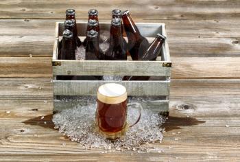 Front view of a frosty large glass mug of dark beer with vintage crate filled with bottled beer and crushed ice on rustic wooden boards. 