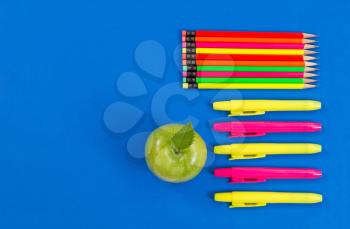 Office or back to school supplies consisting of a green apple, highlight markers and colorful pencils on blue background.  
