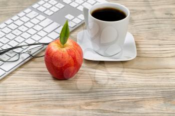 Close up of a red apple with computer keyboard, reading glasses and cup of coffee in background on desktop. 