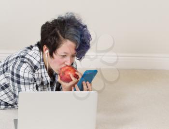 Teen girl eating apple while looking at cell phone with computer in forefront while lying down listening to music at home. 