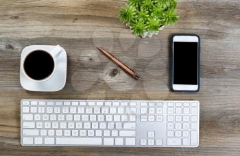 Top view of a neat desktop with keyboard, black coffee, green plant and cell phone on wooden desk.