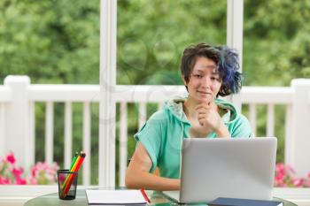 Smiling teen girl, looking forward, with computer, books and pencils in forefront. Large windows in background with blurred out bright green trees and flowers.  