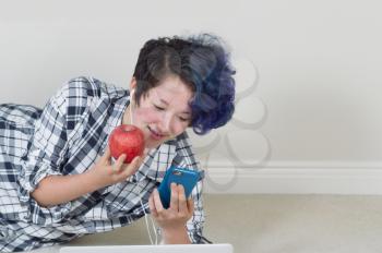 Teen girl holding apple while looking at cell phone with computer in forefront while lying down listening to music at home. 