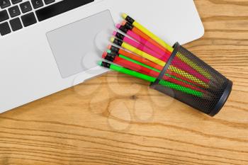 Top view of partial computer laptop with colorful pencils in cup on desktop. 