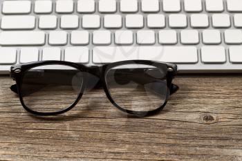 Selective focus on reading glasses with partial keyboard in background. Layout in horizontal format on rustic wood. 