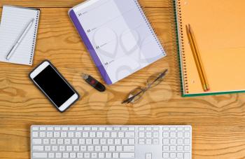 High angled view of desktop consisting of computer keyboard, pencils, pen, calendar, reading glasses, cell phone, notepad and thumb drive. Horizontal layout. 