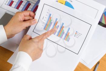 Woman hand pointing index finger at bar chart with printed graphs in background on desktop.