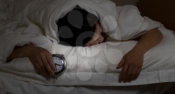 Woman with insomnia, under blanket looking down, holding alarm clock in hand. Select light and focus on woman and clock with darker background for night time concept.