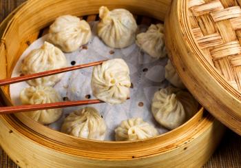 Close up of Chinese steamed dumplings, selective focus on piece held in chopsticks, being taken out of bamboo steamer with rustic wood in background.   