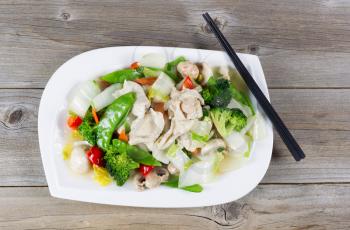 Top view of stir fried white chicken pieces with broccoli, snow peas, peppers and mushroom in white plate on rustic wood setting. 