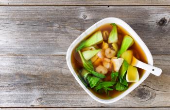 Top view Chinese soup dish with shrimp, fish, bok choy, and spoon on rustic wood. 