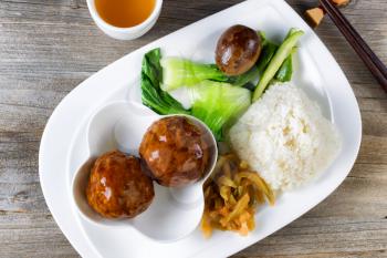 High angled view of Asian saucy meatballs, rice, egg, cucumber and bok choy on white plate. Chopsticks and green tea in background. 