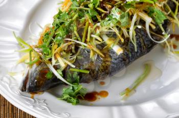 Close up a freshly steamed whole fish covered with herbs, onions and sauce on white plate.