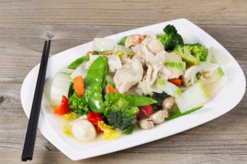 Close up view of stir fried white chicken pieces with broccoli, snow peas, peppers and mushroom in white plate on rustic wood setting. 