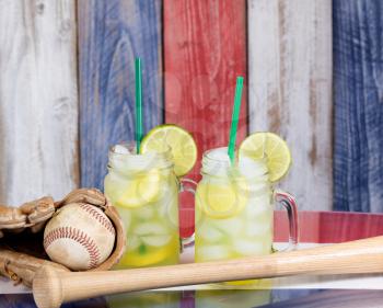 Jar glasses filled with cold lemonade with baseball sporting items.  Faded wooden boards painted red, white and blue in background. Selective focus on upper front jar glass with lime slice. 