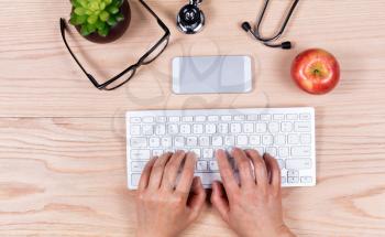 Overhead view of female hands typing on computer keyboard with medical items on wooden desktop. 