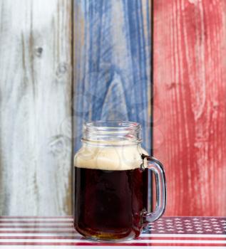 Front view of a single jar glass of cold dark beer.  Faded wooden boards painted red, white and blue in background with USA flag underneath.  