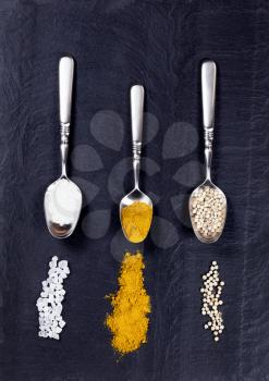 Overhead view of three old spoons containing various spices with trails on natural black slate stone. 