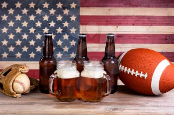 Two pint jars filled with beer, full bottles, football and baseball mitt with vintage wooden USA flag in background.