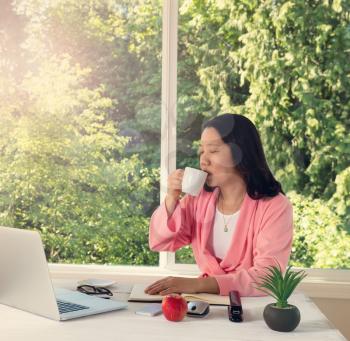 Mature woman, eyes closed, enjoying her morning coffee while working from home in front of large daylight window. Light haze effect applied to image. 