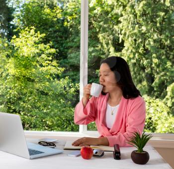 Mature woman, eyes closed, enjoying her morning coffee while working from home in front of large daylight window. 