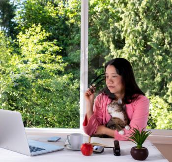 Mature woman, wearing pink bathrobe, holding her family cat while working from home in front of large window with bright daylight and trees in background. 
