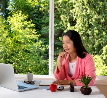 Mature woman, wearing pink bathrobe, working from home in front of large window with bright daylight and trees in background. 