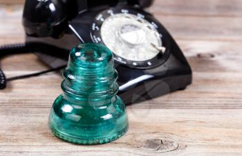 Close up of an antique electric glass insulator with vintage manual dial phone in background. Rustic wood underneath telecommunication objects.  