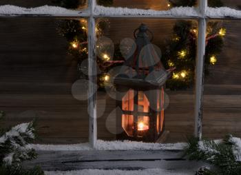 View of a glowing lantern and Christmas wreath through a snow covered window with fir branches.