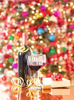 Wine glasses, vintage cork screw, present, and unopen wine bottle on Mahoney table with bright Christmas tree lights in background.  Vertical format layout. 
