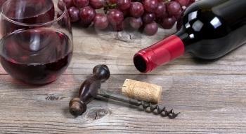 Selective focus on vintage corkscrew with a bottle of red wine, grapes, and drinking glasses in background on rustic wooden boards