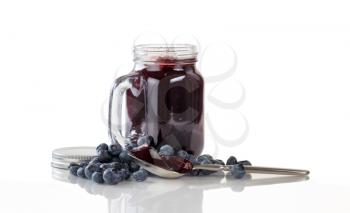 Blueberry jam in spoon with fresh ripe blueberries and jar in background. Isolated on white with reflection.