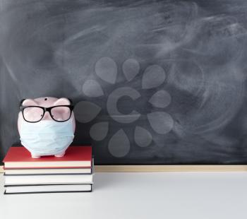 Piggy bank with surgical mask and reading glasses on face with chalkboard in background for copy space 
