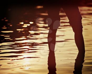 Female legs in rolled jeans in sea water on soft background of setting sun with retro effect filter.