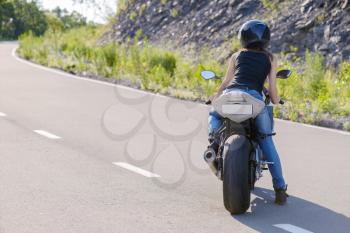 Young beautiful blonde girl in trendy blue jeans and a black t-shirt rides on modern motorcycle.  Outdoor portrait in soft sunny colors.
