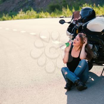 Young beautiful blonde girl in trendy jeans and a black t-shirt sits and drinks water near the modern motorcycle. Outdoor portrait in soft sunlight.