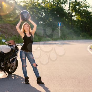 Young beautiful blonde girl in trendy jeans and a black t-shirt removes his helmet near modern motorcycle. Outdoor portrait in soft sunny colors.
