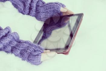 Woman hands in light teal knitted mittens are holding modern tablet pc on snow background. Winter concept.