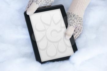 Woman hands in light teal knitted mittens are holding modern tablet pc with blanc screen on snow background. Winter concept.