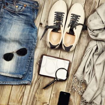 Set of travel items including scarf, sunglasses, sneakers, note book, magnifying glass, mobile phone and jeans. Different travel objects and passenger luggage on grey wooden background. Top aerial vie