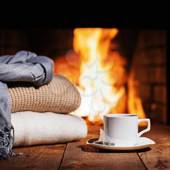 White cup of tea and warm woolen things near fireplace on wooden table. Winter and Christmas holiday concept.