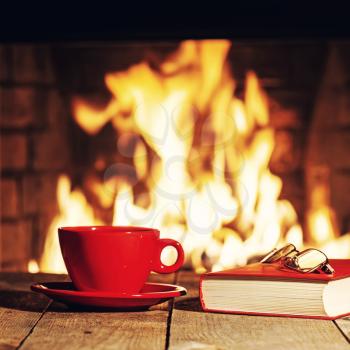 Red cup of tea or coffee, glasses and old book near fireplace on wooden table. Winter and Christmas holiday concept. Photo with retro filter effect.